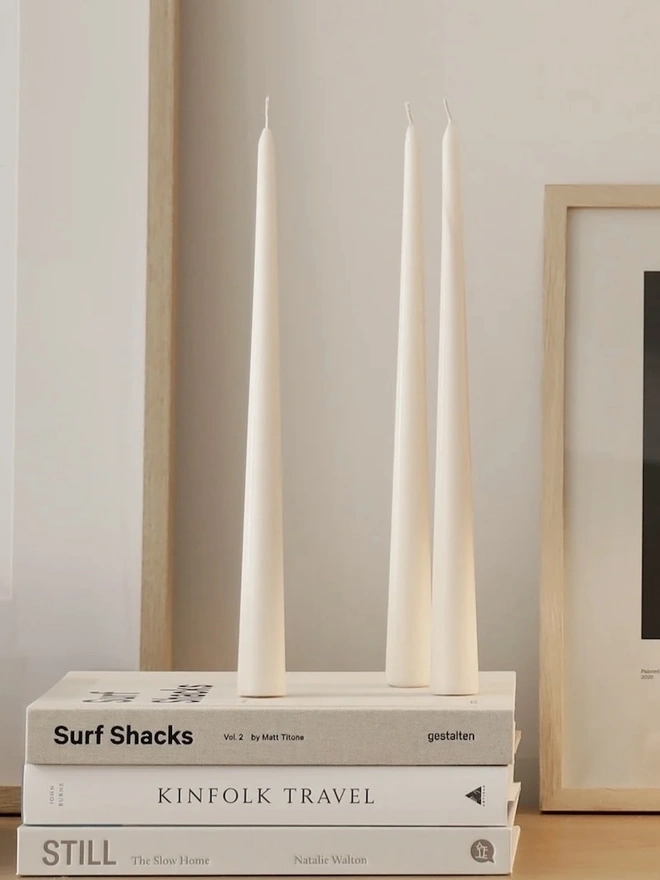 White Stem Candles (Set Of 3). Three unlit cream taper candles balanced on a stack of neutral books in front of a light wood frame. The sleek freestanding shape needs no holder, which makes it easy to style on shelves and sideboards.