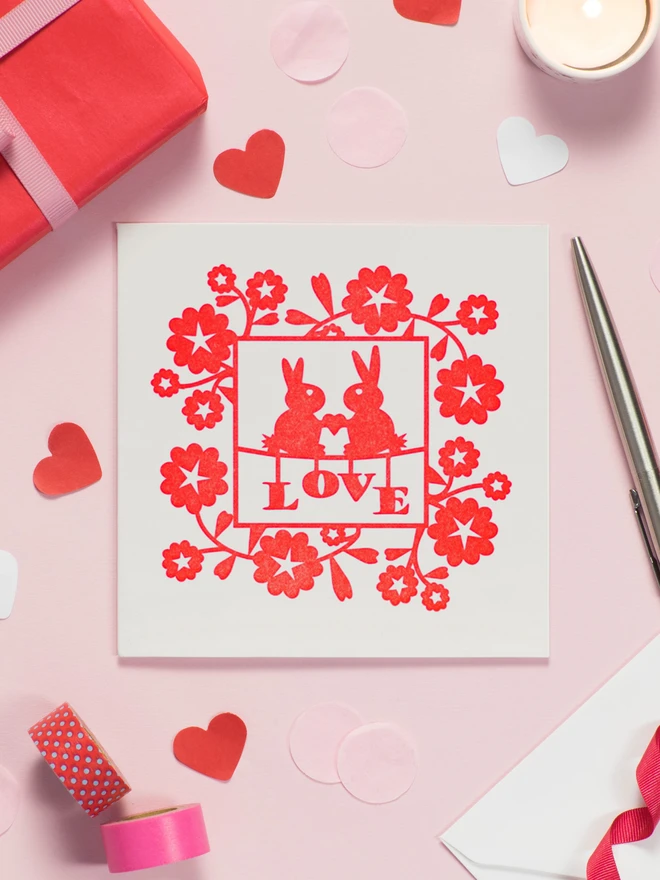 Square Off White Card featuring letterpress deisgn of two rabbits holding a heart between them above the word Love surrounded by a square border of red flowers