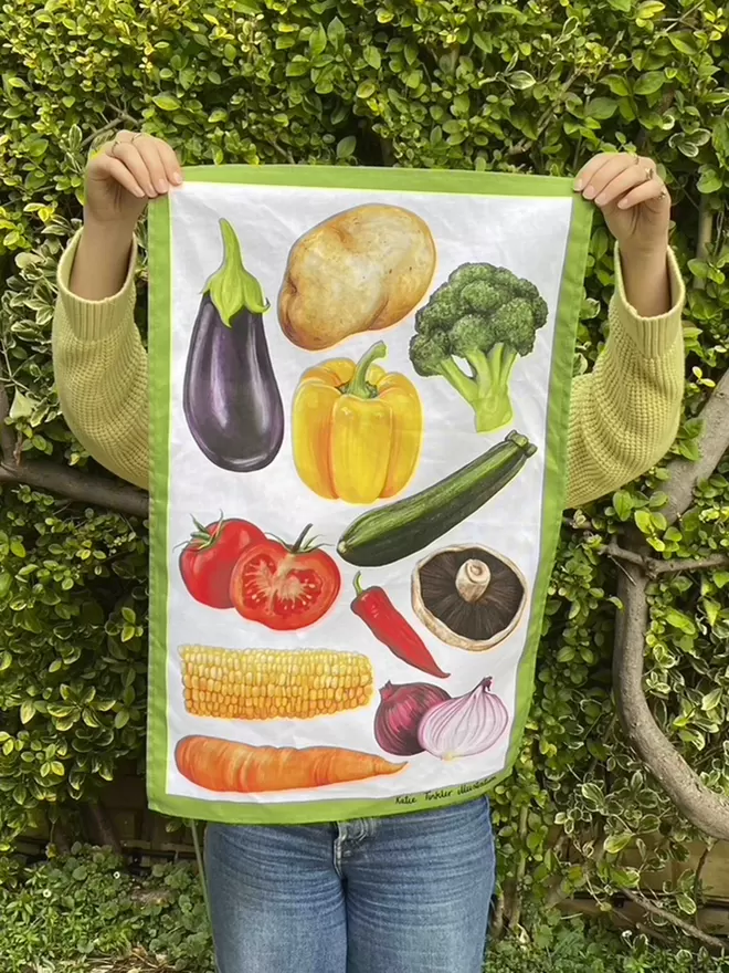A vegetable themed tea towel being held by someone stood in a garden