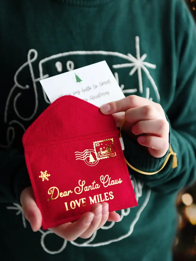 A child wearing a deep green jumper is holding a handmade red felt Christmas envelope that reads "Dear Santa Claus, Love Miles," with a festive letter card inside.