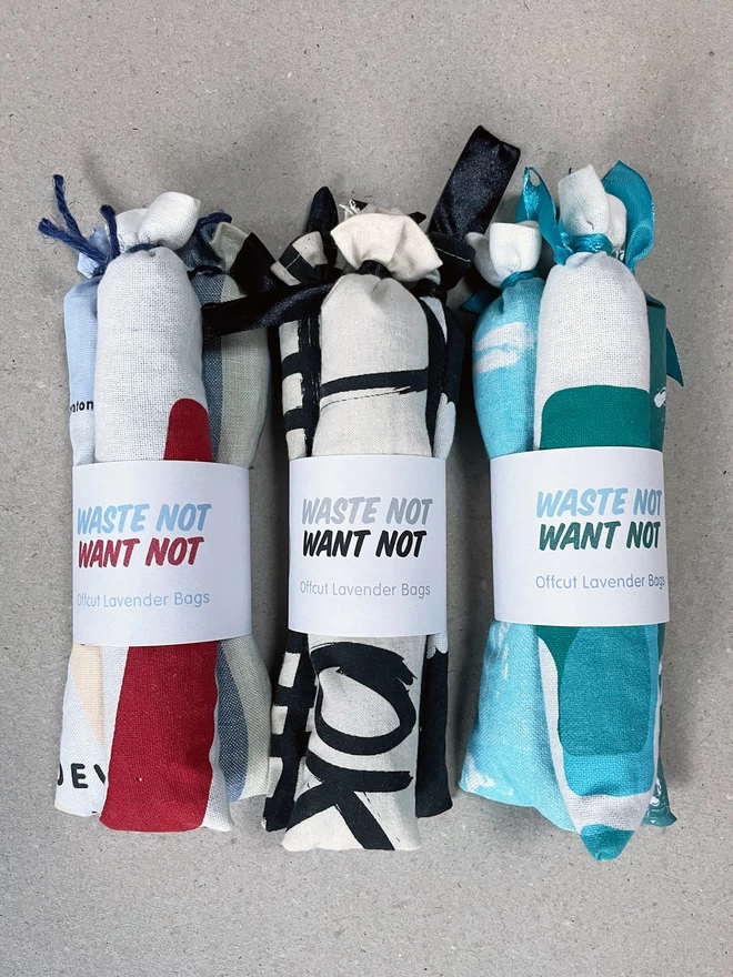 Three sets of three long style cotton lavender bags, tied at the tops, gathered with a branded cuff, sit on a greyboard background - it says Waste Not Want Not, Offcut Lavender Bags on the label. Each bundle displays a different colour way, here is a red, blue and cream - then a black and white - and the turquoise and teal patterns.