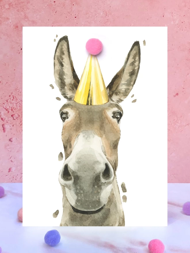 A greeting card featuring a hand painted design of a donkey, stood upright on a marble surface surrounded by pompoms. 