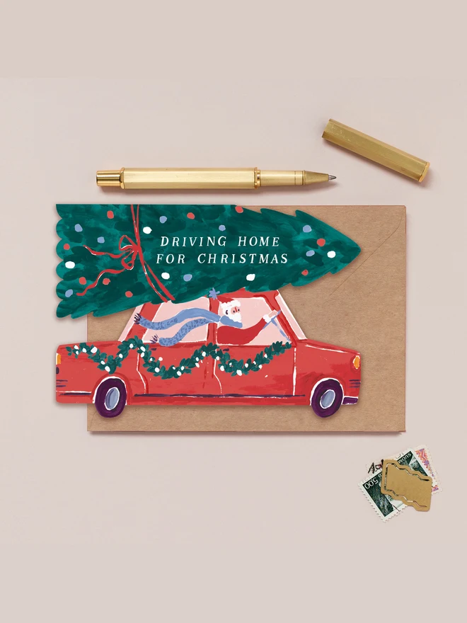 Driving Home For Christmas card shaped like Santa driving a red retro car with a Christmas tree on the roof, on a kraft envelope beside a gold pen