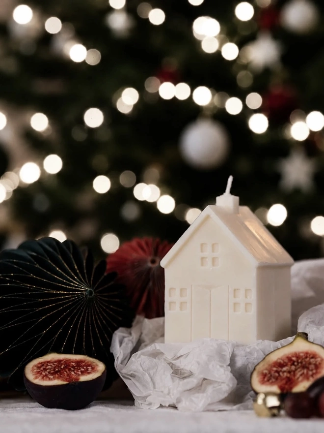 White Christmas Townhouse Candles (Set Of 3). A small cream house candle with a chimney wick, nestled in white tissue beside halved figs and paper honeycomb baubles in front of a lit Christmas tree. The charming festive candle makes a lovely stocking filler or Secret Santa gift.