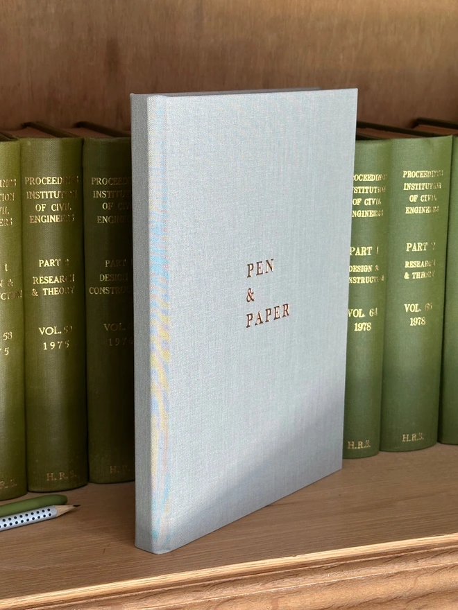 A grey pale grey notebook is propped up on a shelf amongst lots of old green books. Looks like a scene from an old fashioned library.