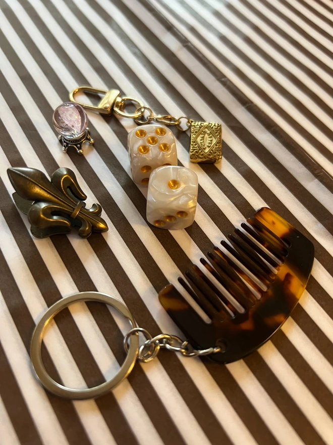 Example of trinkets found in cracker, including miniature tortoiseshell comb keyring, golden dice, custard cream keyring, fleur de lid notebook clip, crystal ball trinket, laid on a striped brown and white background 