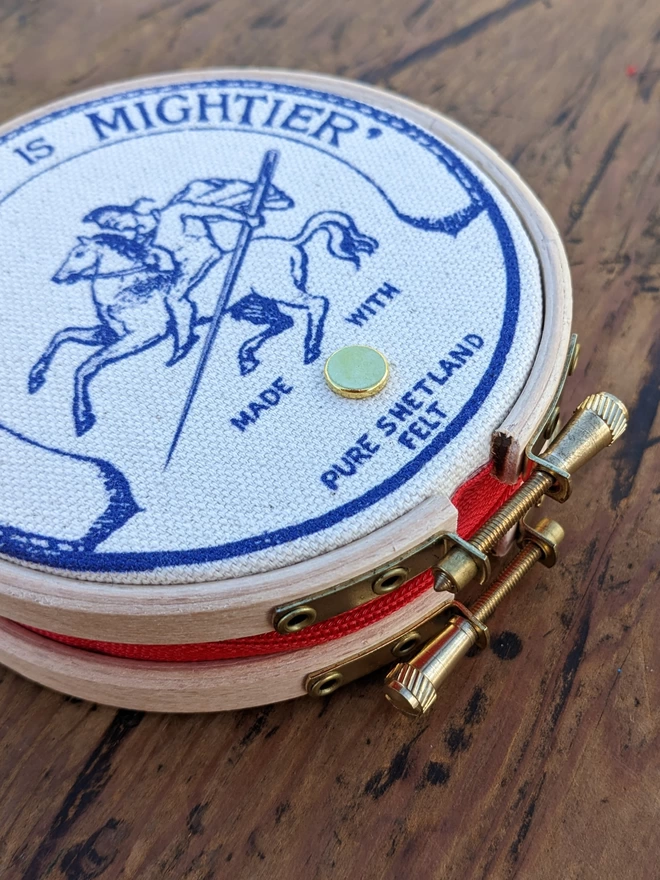 close up of royal blue coloured 'the needle is mightier' embroidery hoop needle book depicting a soldier on horse back with a needle as a sword