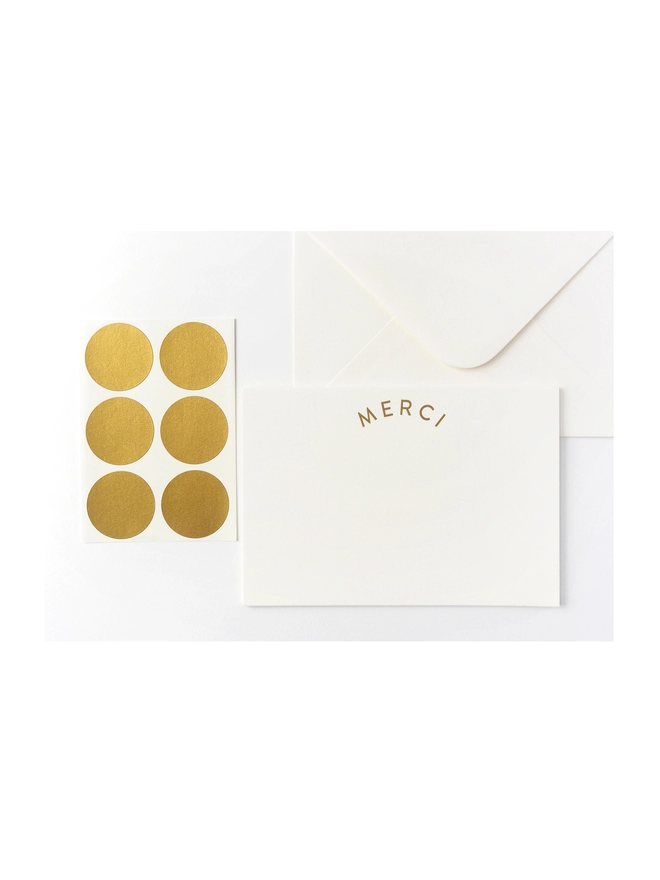 Notecard on white paper that says 'Merci' in Gold Foil, with an Envelope that has a golden seal