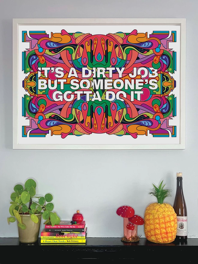 The bold print with the words "It's a dirty job but someone's gotta do it" is hung in a white frame above a shelf decorated with a pot plant, books, a toadstool ornament, a plastic pineapple and a wine bottle.