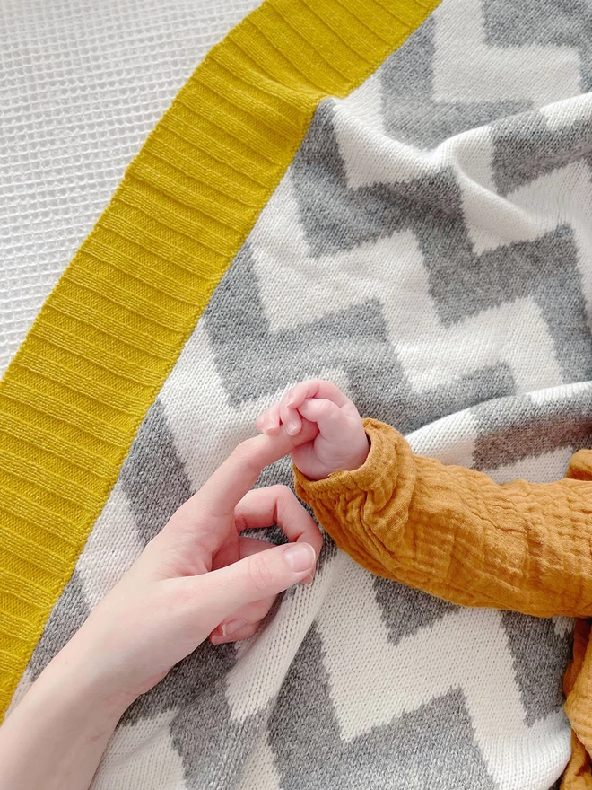An overhead view of the corner of a grey and white zigzag knitted blanket with a bright mustard ribbed trim. A tiny baby hand is visible holding an adult hand.