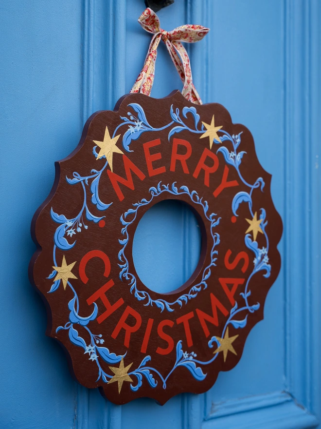 Depp Reddish Brown Hand painted Christmas Wreath with red lettering and gold leaf stars on a blue front door