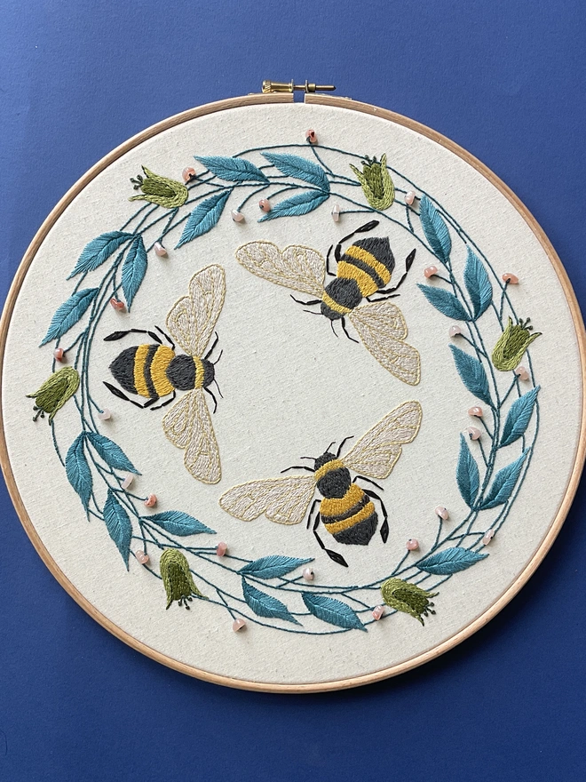Embroidery hoop containing hand stitched bees blue leaves green flowers coral coloured beads