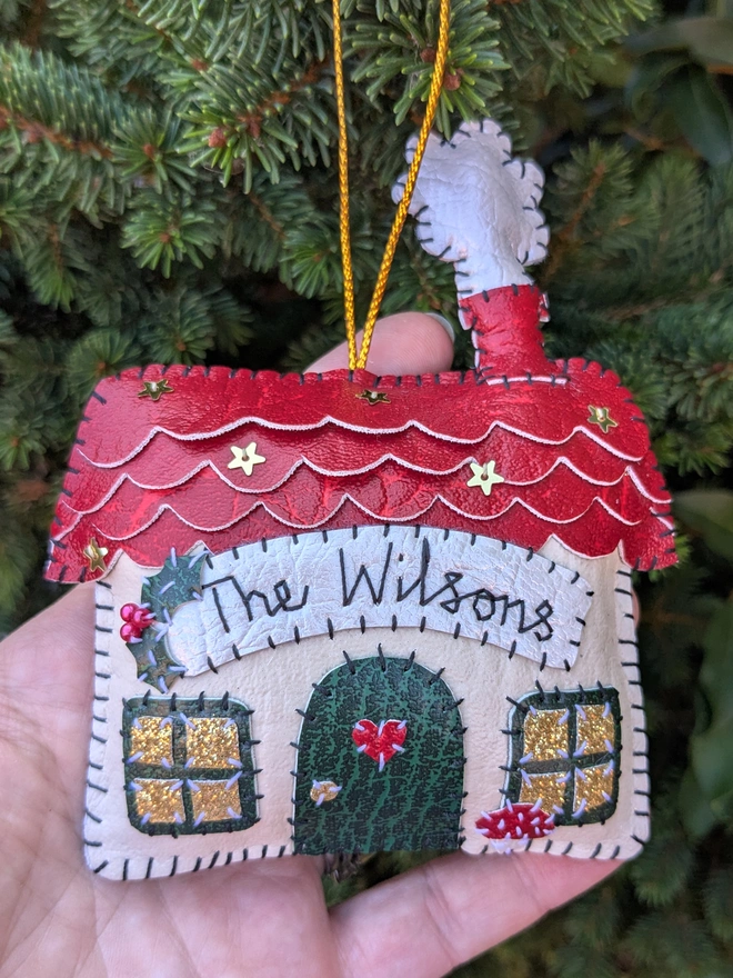 Hand stitched personalised Christmas house decoration made from vegan-friendly faux leather, featuring a red roof, gold stars and a family name banner.