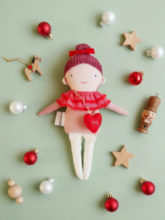 Handmade cloth doll with a pink corduroy hair with a red velvet bow and a layered red ruffle collar and pink outfit, wearing a small red felt heart bag with the letter “M”