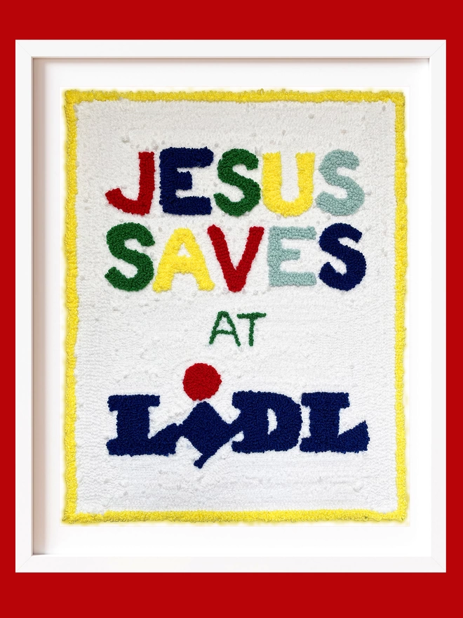 textile artwork with Jesus saves at Lidl written on a white background in bright coloured wool in a white box frame on a red background
