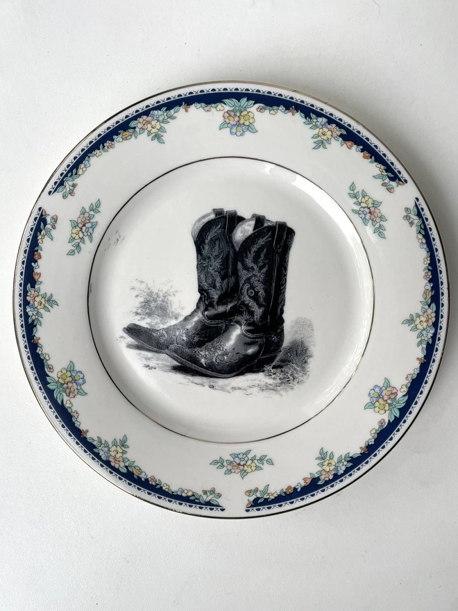a vintage plate with an ornate border has a black and white vintage illustration of a pair of ornate cowboy boots in the centre