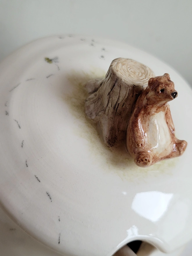 close up of a ceramic white honey jar with a brown bear sitting against a tree stump with a tiny bee and paw prints