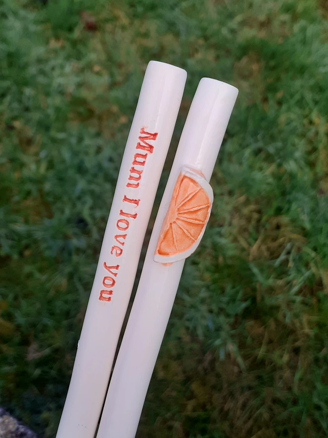 Two white ceramic drinking straws photographed outdoors on grass, one decorated with a raised orange slice motif and the other reading “Mum I love you” in red lettering.