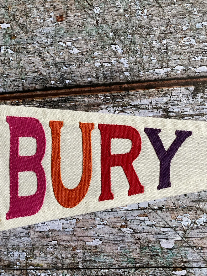 Detail of an ivory canvas Glatonbury pennant flag with the letters BURY in pink, orange, red and purple canvas.