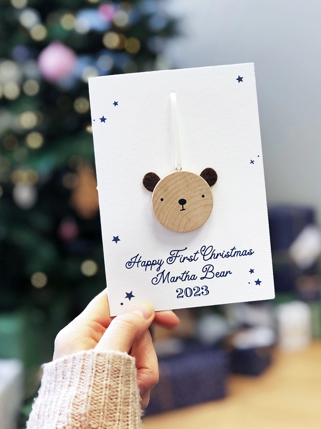 A first Christmas greetings card with a small wooden bear decoration attached is being held in front of a Christmas tree.