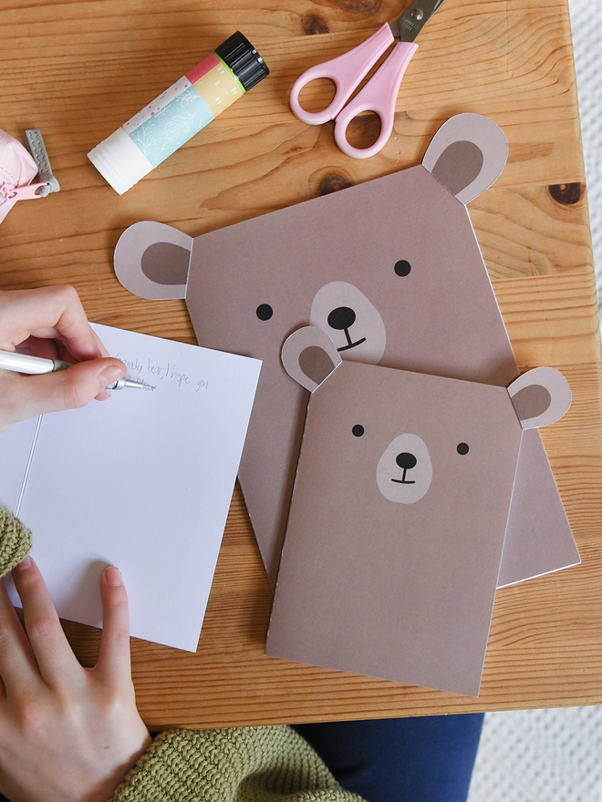 Several brown bear greetings cards lay on a wooden table. A child is writing a message in one of the cards.