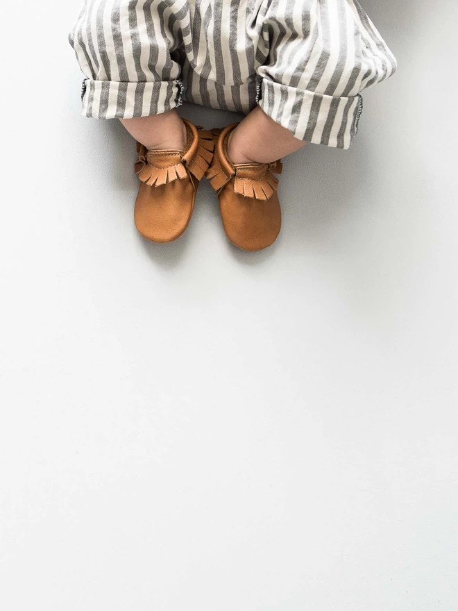 A small baby wearing classic tan Amy and Ivor baby moccasins.