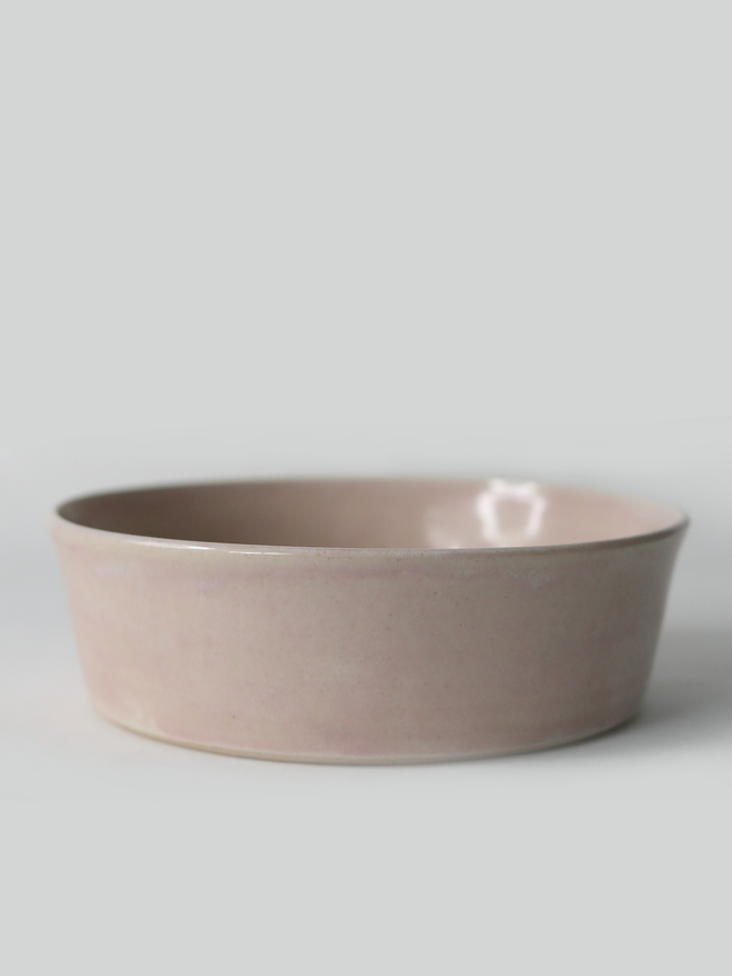 A single wide, shallow glazed ceramic pasta bowl in blush pink, photographed from a slight angle against a plain light grey background, showing the unglazed natural clay base.