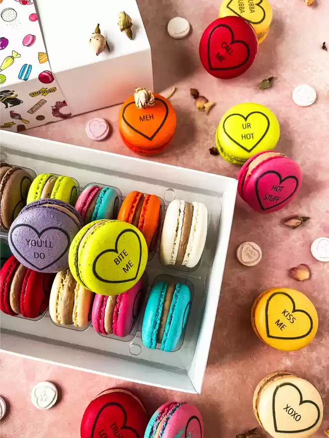 Colourful macarons decorated like conversation love heart candy in a white gift box on a pink background with colourful roses
