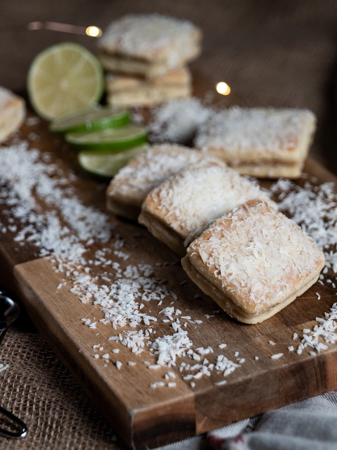 Lime and coconut welshcakes on a wooden chopping board surrounded by desiccated coconut and sliced lime 