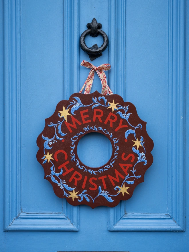 Depp Reddish Brown Hand painted Christmas Wreath with red lettering and gold leaf stars on a blue front door