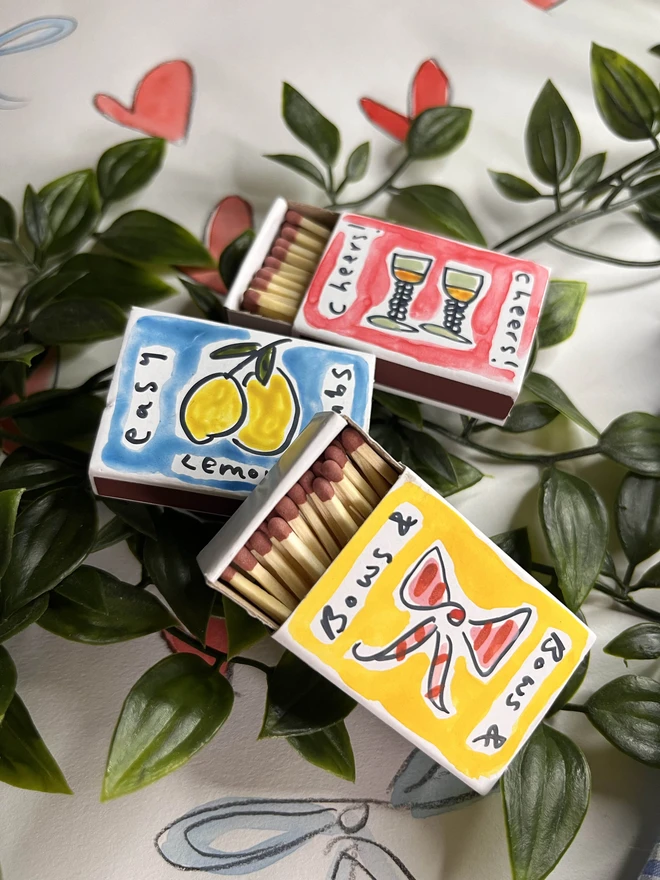 A collection of three hand-painted matchboxes with playful and colorful designs: one features two glasses with the word "Cheers!" written in bold lettering, another displays bright yellow lemons with "Easy Squeezy" written alongside, and the third showcases a red bow with the phrase "Bows & Arrows." Each matchbox is crafted with vibrant illustrations and creative lettering.