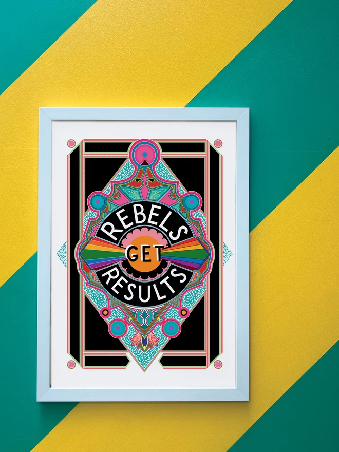 Rebels Get Results is written in white on a black background at the centre of this vibrant, abstract portrait illustration, with a black background and rainbows emitting from the centre, and blue and pink detailing.. The picture is hanging in a white portrait frame against a wall painted with thick diagonal green and yellow stripes.