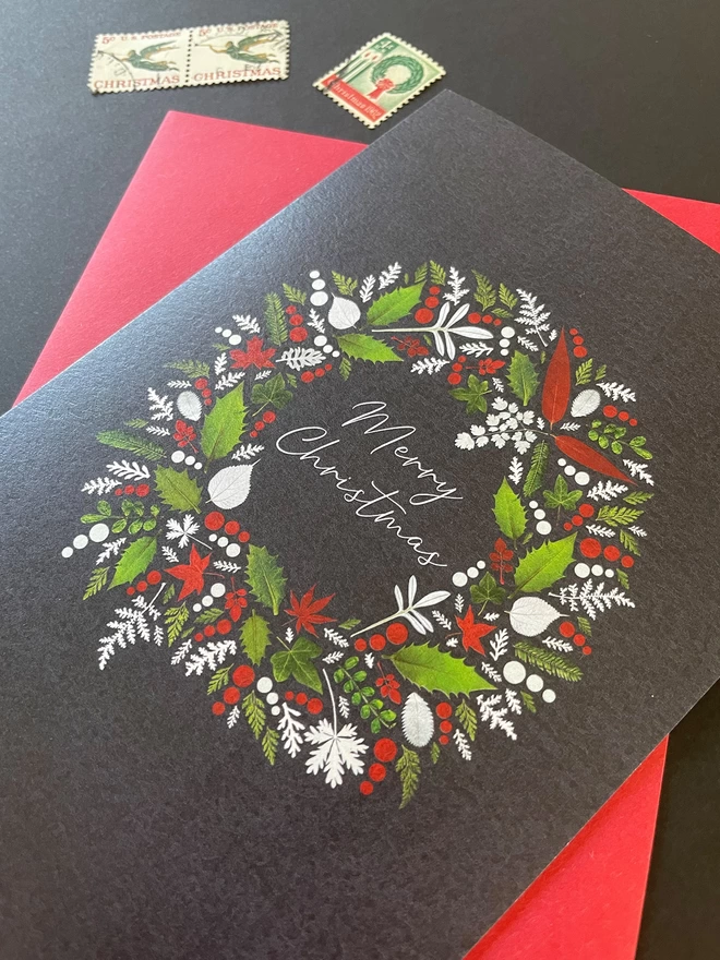 Close-Up of Christmas Card with Pressed Winter Leaves Wreath Design, Holly, Ivy, and Red Envelope on Dark Charcoal Desk