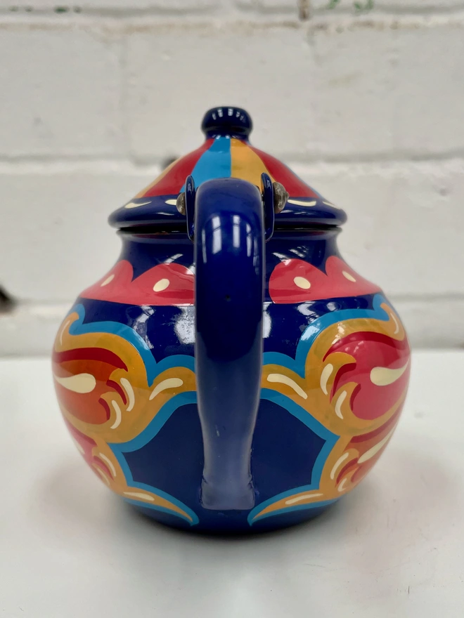 Small blue vintage enamel tea pot, hand painted with a fairground style design in pink, orange, and red. Teapot has a hinged lid.