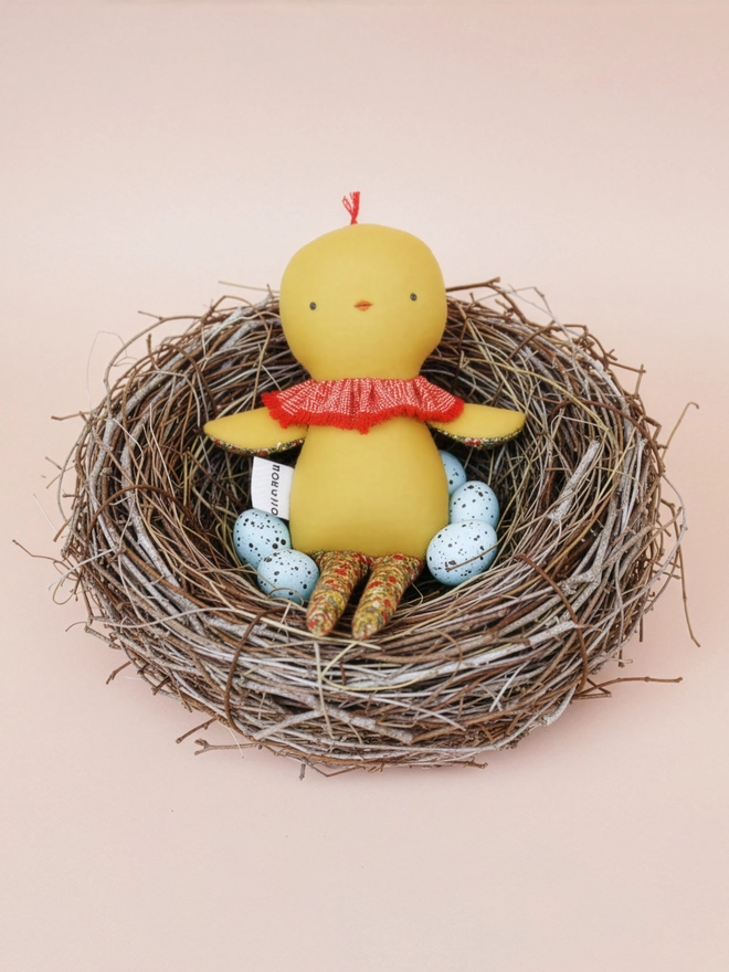 Handmade chick doll made from soft yellow fabric, with a stitched face, small beak, ruffled collar, and patterned fabric arms and long legs, sat in a nest made with twigs, with speckled eggs.