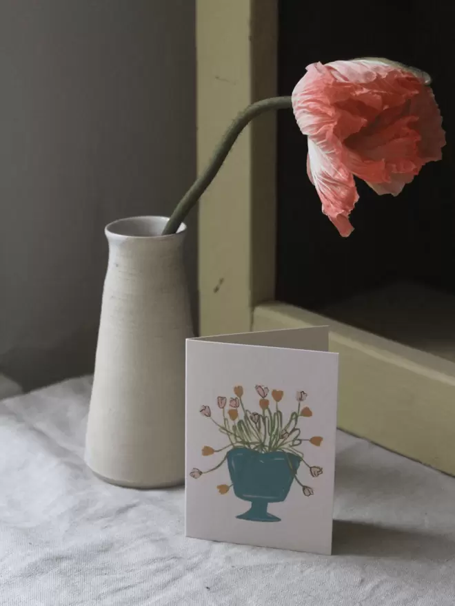 card with tulips arranged in a bright blue vase, next to yellow shelf and icelandic poppy in a cream vase. 