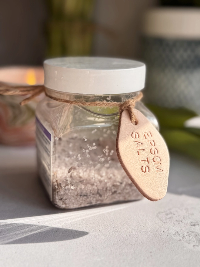 hand-stamped leather tags in natural leather, with the words  'EPSOM SALTS,' stamped onto the tag. Each tag has a cord for tying around jars, displayed on a light, textured surface.