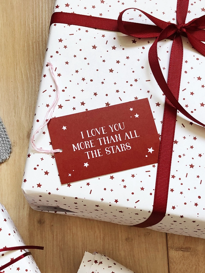 A gift wrapped in white wrapping paper with a red star design, tied with red ribbon, and a tag that reads "I love you more than all the stars" is on a wooden surface.