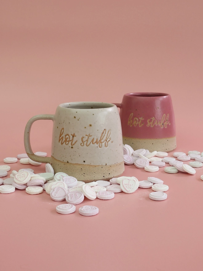 two handmade ceramic mugs in pink and white with the phrase hot stuff on the front sit on a pink background surrounded by love heart sweets 