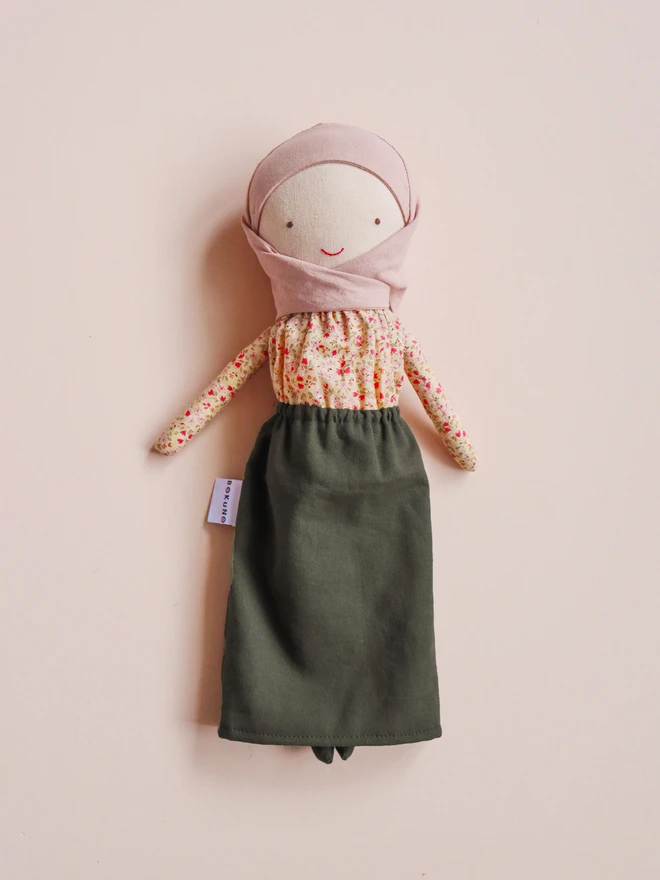 Handmade cloth doll wearing a blush pink hijab, a beige floral long-sleeve blouse, and an olive green skirt. The doll has light fabric skin, simple stitched eyes and a small red smile, with a BOKUNO label on the side. Photographed against a plain light background.