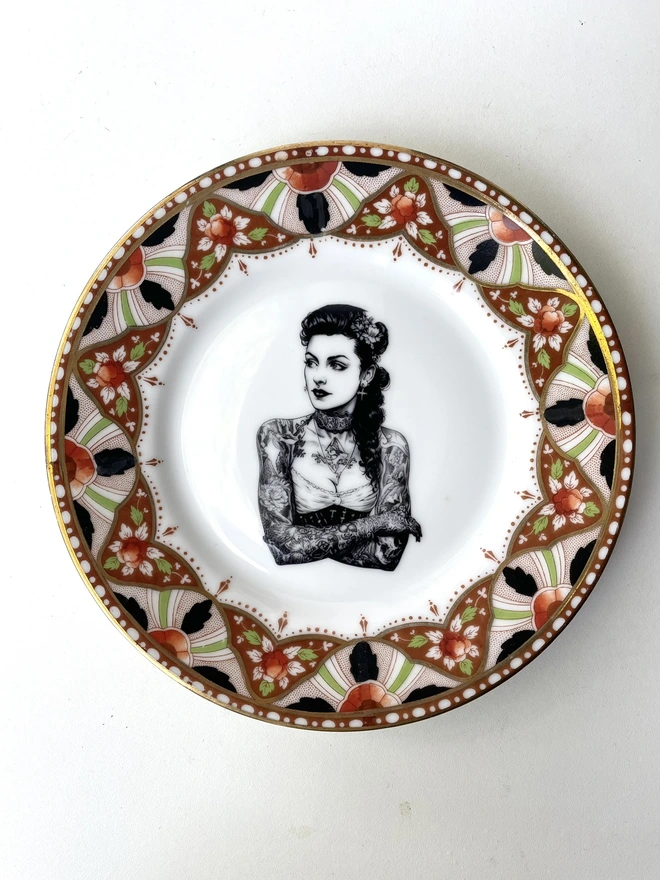 a beautifully ornate plate and in the centre is a vintage image of a beautiful tattooed london lady, she has ringlets and a choker and a tattooed ship on her chest
