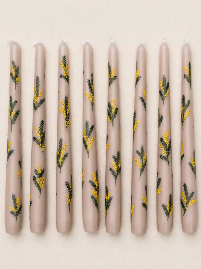 Eight hand-painted oat-coloured taper candles with delicate yellow mimosa flowers and green foliage, arranged in a neat row on natural linen fabric.