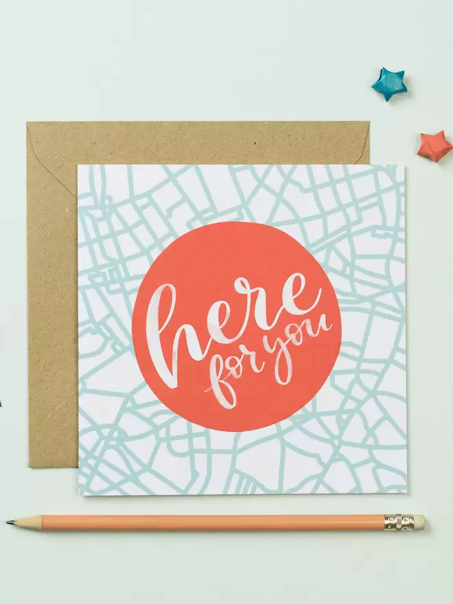 A square card features a red circle that says 'here for you' in white lettering. The background of the card is an abstract map design in duck egg blue. Tucked beneath it is a brown envelope.