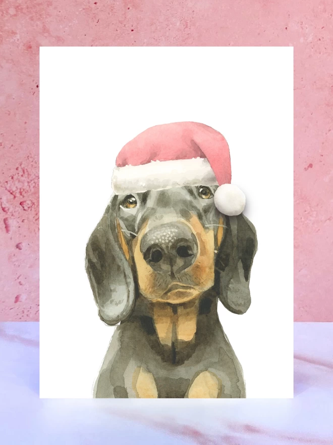 A Christmas card featuring a hand painted design of a black and tan dachshund, stood upright on a marble surface surrounded by pompoms. 