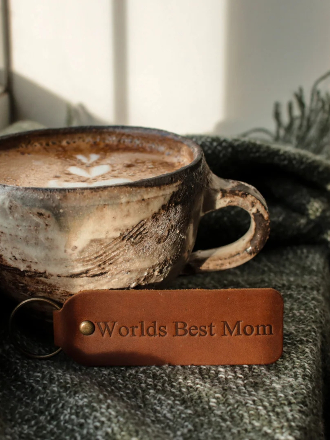 mother's day leather keyring. a brown leather keyring propped up against a coffee cup, embossed with 'world's best mom'.