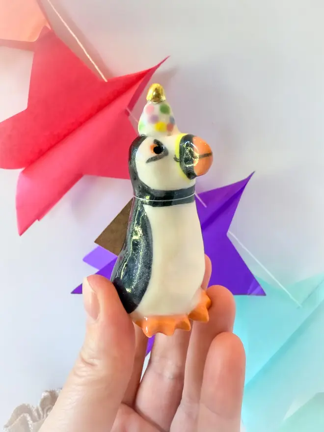 Handmade Porcelain Puffin Penguin Topper With Golden Hat, held by hand near party decorations. 