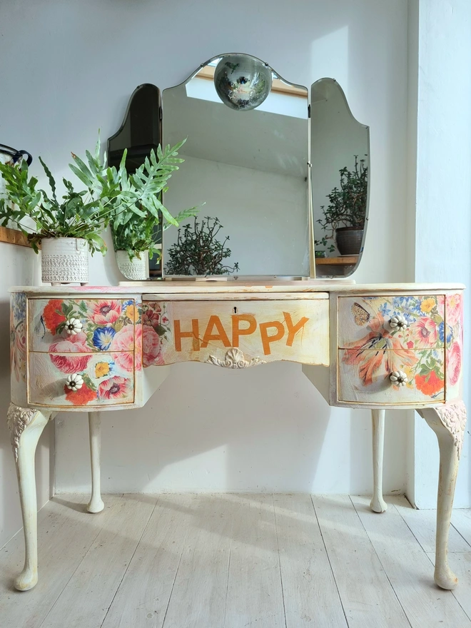 An antique kidney-shaped dressing table with chair and a trio of mirrors. Upcycled in cream and pale pink with decoupage florals in oranges and pinks. Text across the table read ‘flowers make me happy’ in the orange of the undercoat.