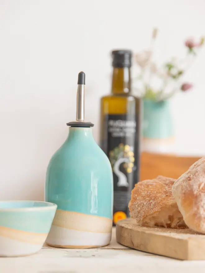 Shoreline oil pourer in turquoise and pink glaze. The bottle has a steel and rubber funnel in the top. Next to the bottle is a matching mini bowl, a chopping board with bread and in the background is a bottle of olive oil and a vase of wildflowers out of focus.