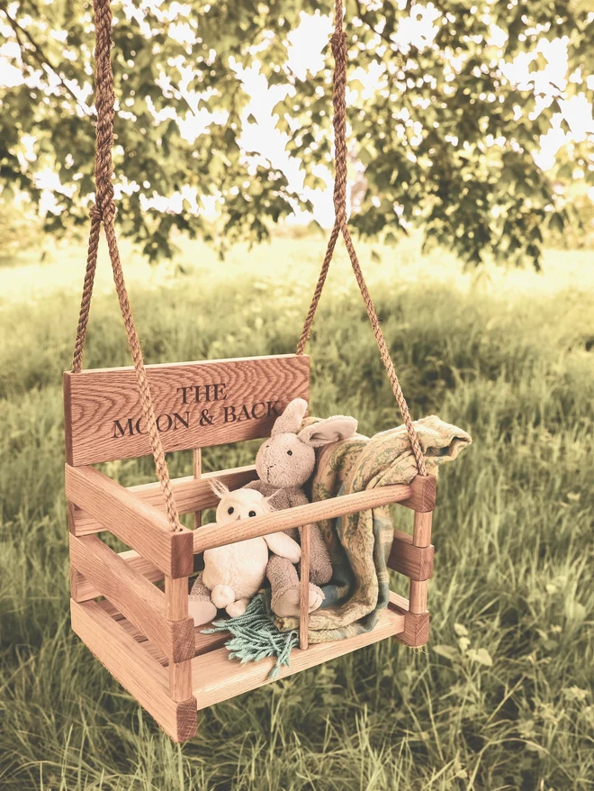 Personalised oak baby swing engraved 'The Moon & Back' with a bunny, owl and blanket tucked inside, hanging outdoors in dappled summer light.