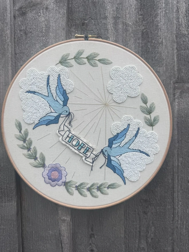 An embroidery hoop containing embroidered blue swallows carrying a banner with the word Hope on, beaded white clouds, gold sun rays and olive leaves surrounding 